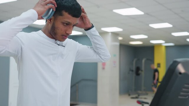 student approaches treadmill clips on over ear headphones powers machine and waits as display boots up in bright fitness studio with stairwell and other exerciser blurred in background