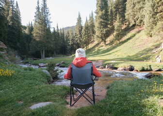 A girl or woman in a red jacket and white hat sits on a tourist chair and admires a mountain summer...