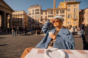 A woman in a blue coat enjoys her morning coffee at a table in front of the Pantheon in Rome,...