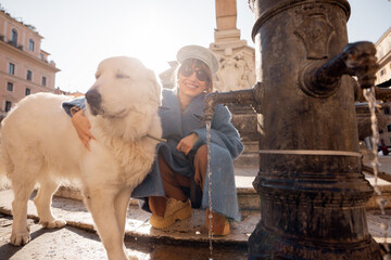A fluffy white dog drinks from a traditional Roman fountain while its owner smiles nearby, with the iconic Pantheon in the sunlit background