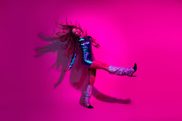Energetic young woman in a glossy blue dress dancing against a vibrant neon pink background