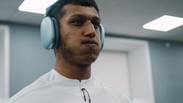 close up of focused man wearing over ear headset and white zip-up top puffing cheeks while exerting visible pressure during barbell lift in well lit gym with blurry door and ceiling lights behind