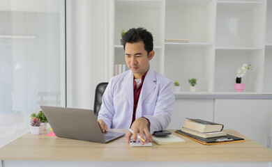 A man in a white lab coat is sitting at a desk with a laptop and a tablet. He is using the tablet to take notes while looking at the laptop. Concept of professionalism and focus