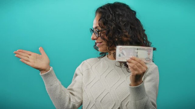 Woman pointing while holding a uae dirham banknote against a blue background, wearing a sweater and glasses, conveying financial concepts with a gesturing hand.