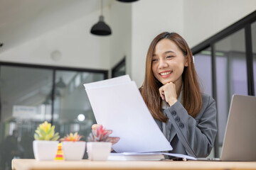 Obraz premium A woman is sitting working in an office. She is at a desk with a laptop and a calculator. She is wearing a gray suit and a white shirt. She is smiling and happy.