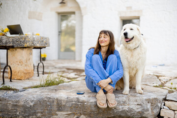 A woman in a blue outfit sits on stone steps beside her large white dog in front of a traditional whitewashed house. The image conveys warmth, trust and the serenity of countryside life