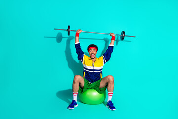 Young man in retro activewear lifting a barbell while seated on a fitness ball in a bright teal...