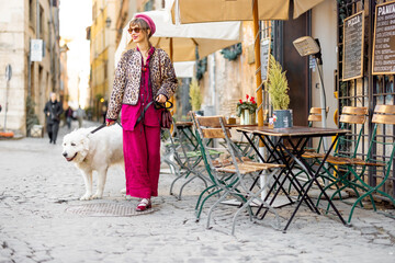 A stylish woman in bold colors walks her fluffy white dog past a charming Roman cafe. The scene captures a relaxed urban stroll and the elegance of everyday life in Italy