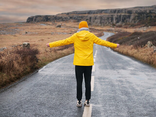 A woman in a yellow jacket walks down a road. The road is wet and the sky is cloudy. Beautiful and wild scenery of Burren area in the background. Travel and tourism in Ireland. Irish landscape. © mark_gusev