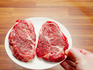Hand holding plate with two fine raw rib eye steaks seasoned with herbs and pepper. Rich expensive beef ready to be cooked. Fine cuts with excellent marbling.