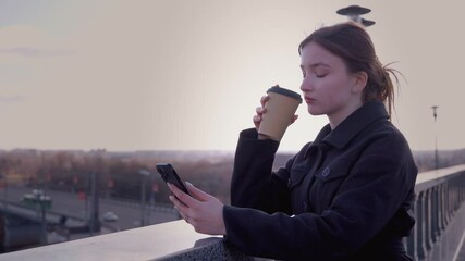 Casually dressed young woman relaxing with takeaway coffee, browsing smartphone while standing on pedestrian bridge, overlooking urban cityscape during golden sunset light