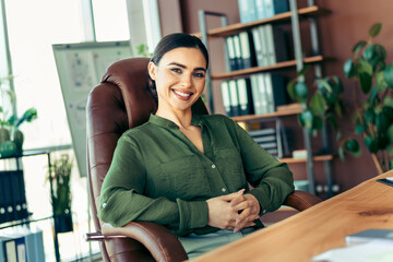 Smiling professional businesswoman in green blouse seated at office desk in a modern workspace,...