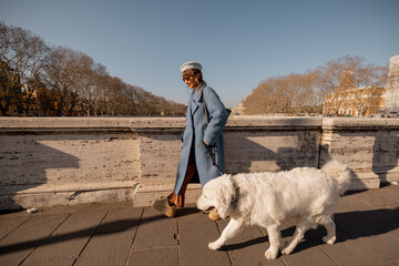 A woman in a blue coat embraces her large white dog on a Roman bridge, gazing toward St. Peters...