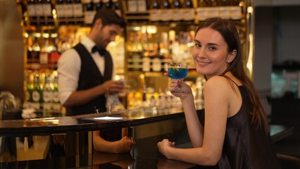Sitting beautiful woman guest looking at camera to pose with drinking mixed vodka in blue hawaii cocktail at luxurious counter bar for Friday meeting on smart bartender background at club. Vinosity.