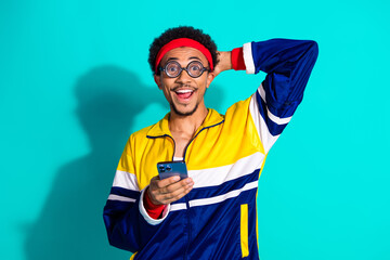 Stylish young man in vibrant retro sportswear smiling joyfully while holding a phone, set against a bold turquoise background