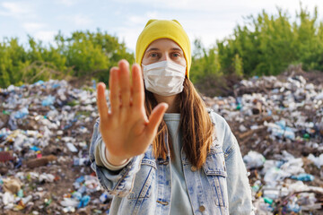 A young woman in a yellow hat and a face mask holds up her hand in front of a landfill, conveying a message to stop pollution.