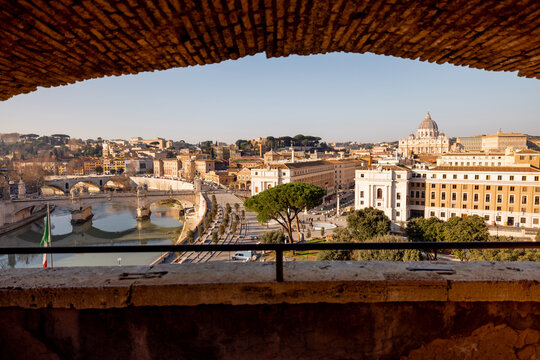 A stunning view of St. Peters Basilica and the Tiber River framed by the brick arch of Castel SantAngelo, capturing the beauty of Rome’s historic skyline in warm morning light - Powered by Adobe