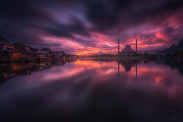Fototapeta premium As the sun sets, vibrant hues of orange and purple reflect on the still lake, showcasing a breathtaking view of a mosque with minarets. The calm water enhances the serene atmosphere.