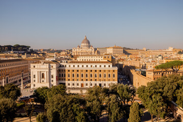 Fototapeta premium Panoramic view of Vatican City and the dome of St. Peters Basilica from the top of Castel Sant Angelo, framed by Roman pines and morning sunlight over the rooftops of Rome