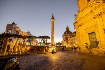 Evening view of Romes iconic Trajans Column and Altare della Patria, illuminated against the twilight sky, surrounded by ruins, churches and cobblestone streets © rh2010