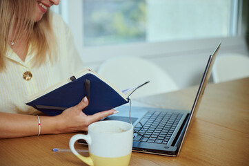 Woman Working Remotely Holding Notebook in Bright Home Office with Laptop and Coffee
