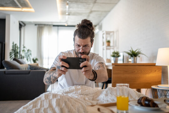 Young man taking photo of breakfast in bed with smartphone