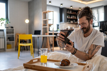 Young man taking photos of breakfast in bed with smartphone