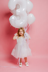 little girl with balloons and the number three on a pink background celebrates her birthday in the studio