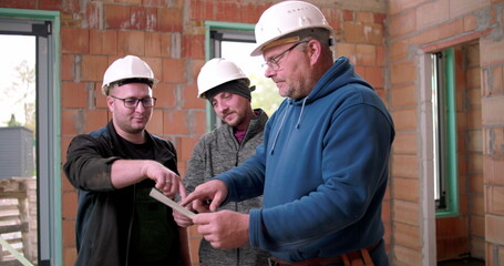 Team of builders reviewing blueprints together indoors, construction planning, three men with...
