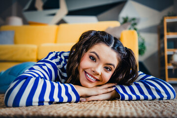Young brunette in striped sweatshirt relaxing on rug in cozy living room with yellow couch and...