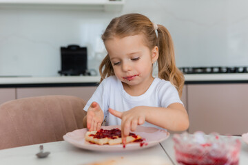 Cute blonde girl eating waffles with jam at home in the kitchen