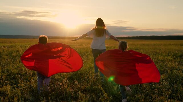 Superheroes Mother, daughter, son playing together on meadow in sunshine. Mom child run in red superhero capes outdoor. Family carnival in sunshine, Halloween. Mom, Super kids dream flying in red cape