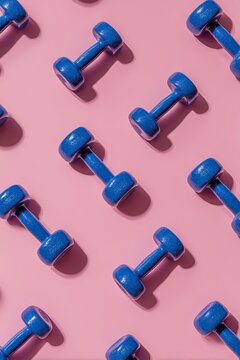 A pink background pattern serves as the backdrop for blue fitness dumbbells, which are essential equipment for home workouts and exercises, as seen in a flat lay gym, symbolizing a healthy lifestyle