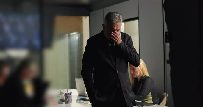 Businessman deeply stressed with hands over face while standing in meeting room as female colleague works in background on laptop during corporate crisis