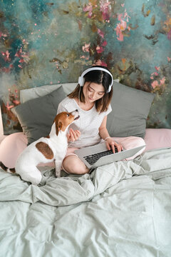 A woman wearing headphones works on her laptop in bed. Her dog sits beside her, looking up, perhaps wanting attention or just keeping her company while she focuses.