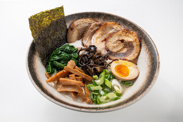 Tonkotsu ramen with noodles, pork belly, seaweed, spinach, mushrooms, egg, and green onions in a rustic brown bowl on a white background. 