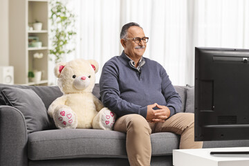 Mature man with a teddy bear sitting on a sofa and watching tv