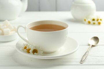 Delicious chamomile tea in cup, fresh flowers, sugar cubes and spoon on white wooden table, closeup