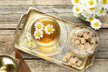 Delicious chamomile tea in glass cup, fresh flowers and brown sugar on wooden table, flat lay