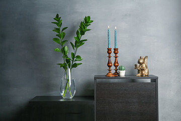 Elegant candlesticks with burning candles, branches in vase and other decor on dresser near grey wall