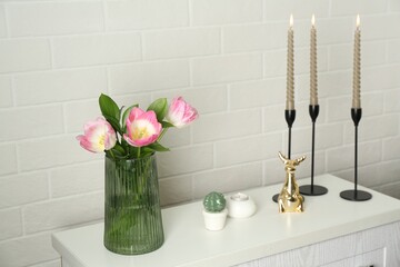 Elegant candlesticks with burning candles, tulips in vase and other decor on dresser near white...
