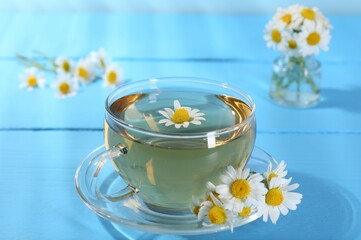 Delicious chamomile tea in glass cup and fresh flowers on light blue wooden table, closeup