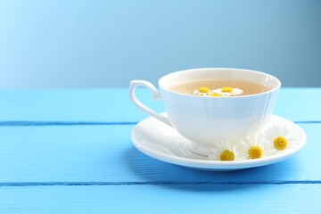 Delicious chamomile tea in cup and fresh flowers on light blue wooden table, closeup. Space for text