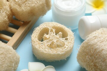 Natural loofah sponges, cosmetic products and plumeria flowers on light blue background, closeup