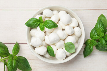 Tasty mozzarella cheese balls in bowl and basil on light wooden table, flat lay