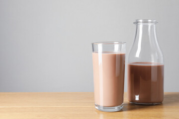 Tasty chocolate milk in glass and bottle on wooden table against grey background, space for text
