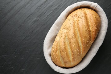 Loaf of bread with sesame seeds in basket on black textured table, top view. Space for text