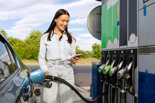 Smiling lady refueling her car and messaging on smartphone at the petrol station, filling up her automobile with bio fuel, pouring petrol into tank of modern vehicle