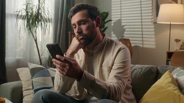 Sad upset frustrated man Caucasian businessman guy bored using mobile phone on couch sofa worried disappointed male homeowner reading news cellphone browsing message scam scrolling smartphone at home