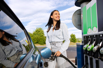 Car fuel offer. Excited woman refilling auto with fuel at modern petrol station, standing near vehicle and looking at gasoline dispenser machine